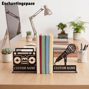 Puede incluir: Un par de sujetalibros de metal negro con un diseño de radiocasete en un lado y un micrófono en el otro. Ambas piezas incluyen una base rectangular con el texto CUSTOM NAME impreso en letras mayúsculas de color blanco.