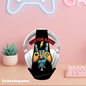 Peut inclure: Un casque blanc repose sur un support noir pour casque avec un graphisme de main de zombie tenant une manette de jeu jaune. Le support porte les inscriptions "CUSTOM NAME" et "Enchantingspace". Un clavier rose et une petite plante succulente sont en arrière-plan.