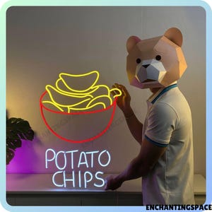 Op de afbeelding: Neon-bord met een gele omtrek van een kom met chips en de tekst "POTATO CHIPS" in wit. Een persoon met een bruin papieren berenmasker staat voor het bord.