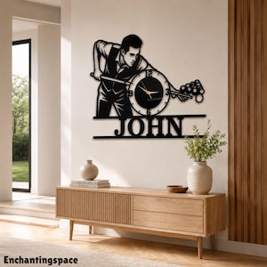 Könnte beinhalten: Eine schwarze Wanduhr mit der Silhouette einer Person, die Billard spielt und ein Queue über einem runden Zifferblatt mit goldenen Zeigern hält. Unter der Uhr ist der Name JOHN in fetten, schwarzen Großbuchstaben als Teil des Designs zu sehen.