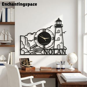 May include: A black metal wall clock featuring a lighthouse silhouette, crashing waves, and rocky cliffs. The circular clock face has a black background with gold hands and the name NOLAN in bold capital letters at the bottom right corner.