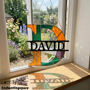 以下が含まれることがあります： 黒い文字「DAVID」が入った、文字「D」の形をしたステンドグラスパネル。オレンジ、緑、紫色のガラスを使用しています。窓の前に吊り下げられており、木製の窓枠に影を落としています。