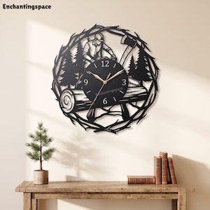 Op de afbeelding: Een zwarte ronde wandklok met een silhouet van een houthakker die met een kettingzaag aan een boomstam werkt, omringd door dennenbomen en een takvormig frame. De klok heeft goudkleurige wijzers en witte cijfers op 3, 6, 9 en 12.
