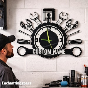 May include: A black and white wall clock shaped like a gear, featuring a central piston, surrounding wrenches and screwdrivers, and a horizontal bar with the text CUSTOM NAME. The clock has a black face with white numbers and bright green hands.