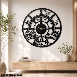 Puede incluir: Un reloj de pared circular negro con diseño de timón de barco, recortes de gaviotas y olas del mar. La esfera central tiene manecillas doradas y los números 3, 6, 9 y 12 en negro. El nombre MAX aparece en letras mayúsculas en la parte inferior.