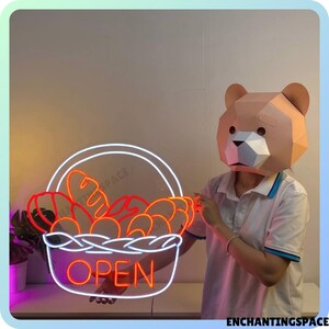 May include: A neon sign shaped like a basket filled with bread, with the word "OPEN" in red neon letters. The sign is held by a person wearing a brown bear mask.