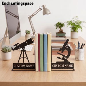 May include: A pair of black metal bookends featuring a telescope silhouette on one side and a microscope silhouette on the other. Each piece has a rectangular base with the text CUSTOM NAME printed in white capital letters on the front.