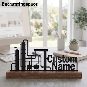 Op de afbeelding: Een zwart metalen bureau-ornament met een silhouet van een industriële raffinaderij met leidingen en tanks, gemonteerd op een rechthoekige houten voet. De tekst Custom Name is in een vet, schreefloos lettertype in het ontwerp verwerkt.
