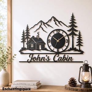 May include: A black metal wall clock featuring a silhouette of a cabin, pine trees, and mountain peaks. The clock face has gold hands and black numerals. Below the clock, the text John's Cabin is cut out in a black script font.