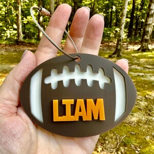 May include: Brown and white football-shaped luggage tag with the name "LIAM" in orange block letters. The tag has a silver-colored wire loop for attachment. The background is a blurred outdoor scene with trees.
