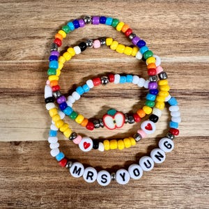 May include: Three beaded bracelets in a rainbow of colors. One bracelet has the text "MRS YOUNG" in white beads. Another has a red heart bead. The third bracelet has an apple bead.
