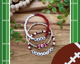 May include: Three beaded bracelets in red, white, and silver. One bracelet spells "BAMA", another has football-shaped beads, and the third spells "ROLLTIDE". The bracelets are on a wooden surface with a football graphic.