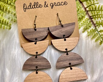 May include: Wooden dangle earrings with a layered, half-circle design. The earrings alternate between dark brown and light brown wood. The earrings are attached to a brown card with the text "fiddle & grace".