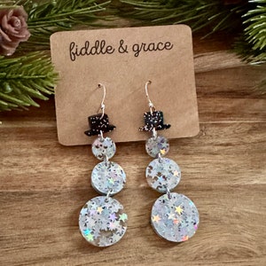 May include: Dangling snowman earrings with silver hooks. Each earring features a black top hat, three graduated, round, glittery sections, and a star-shaped glitter design. The earrings are displayed on a brown tag with the text "fiddle & grace".
