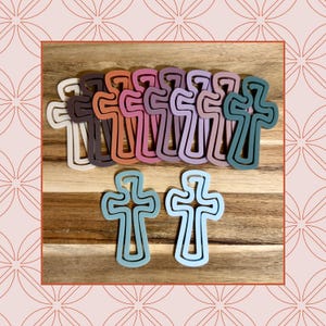 May include: A collection of colorful cross-shaped cutouts in various shades, including white, brown, orange, pink, purple, and teal. Two additional blue-green crosses are positioned at the bottom. The crosses are arranged on a wooden surface.