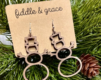 Puede incluir: Pendientes de madera con forma de muñeco de nieve y ganchos plateados. Los pendientes están cortados de madera marrón claro y presentan un diseño de muñeco de nieve con sombrero de copa y brazos extendidos. El texto "fiddle & grace" es visible.