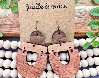 May include: Wooden earrings with a geometric design. The earrings feature a dark brown circular top, a light brown rectangular middle piece, and a reddish-brown U-shaped bottom. The earrings are displayed on a brown card with the text "fiddle & grace".