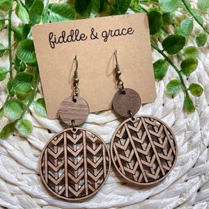May include: Wooden earrings with a geometric design. The earrings have a small, round top piece and a larger, circular bottom piece with a cut-out chevron pattern. The earrings are brown and hang from bronze-colored hooks. The tag reads "fiddle & grace".