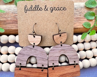 May include: Wooden earrings with a geometric design. The earrings feature a half-circle, an arch, and a rectangle in shades of brown and gray. The earrings are hanging from bronze-colored hooks. The earrings are displayed in front of a brown card with the text "fiddle & grace".