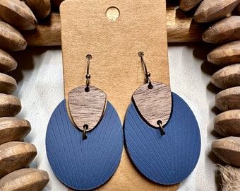 May include: A pair of handcrafted earrings. Each earring features a dark blue, textured, circular base with a chevron pattern, topped with a wooden, geometric accent. The earrings hang from bronze-colored hooks and are displayed on a brown card.