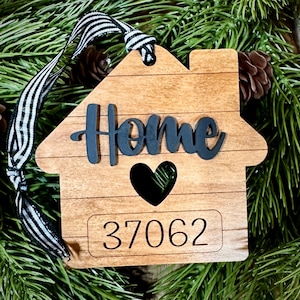May include: Wooden house-shaped ornament with the word "Home" in black script, a black heart cutout, and the number "37062". The ornament has a striped black and white ribbon for hanging. The background includes pine needles and pine cones.
