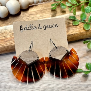 May include: A pair of dangle earrings with a wood grain top and a fan-shaped bottom in shades of brown and amber. The earrings are attached to a brown card with the text "fiddle & grace".