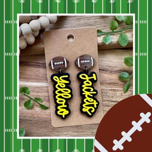 May include: A pair of football-themed earrings. Each earring features a wooden football shape at the top and the word "Yellow Jackets" in yellow script on a black background. The earrings are attached to a brown card.