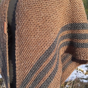 May include: A brown knitted poncho with dark gray stripes on the sleeve. The poncho has a textured appearance and a leather clasp. The background shows a snowy landscape with trees.