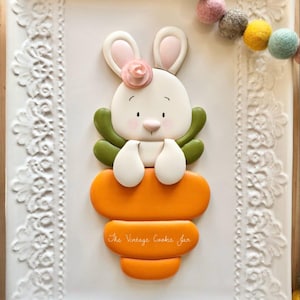 May include: An Easter-themed cookie shaped like a white bunny with a pink flower, holding a carrot. The cookie sits on a white tray with a lace design. The text "The Vintage Cookie Jar" is on the carrot. Pastel felt balls are in the background.