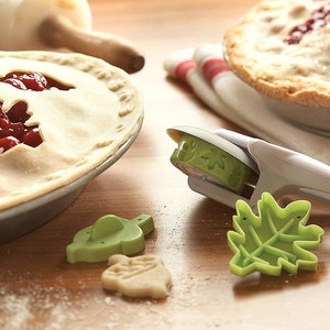 May include: A set of green leaf and acorn shaped cookie cutters, a rolling pin, and two cherry pies. The rolling pin is made of wood and the cookie cutters are made of plastic.