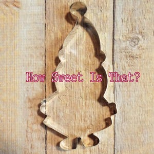 May include: A metal cookie cutter shaped like a cartoon gnome wearing a hat. The cutter is on a wooden surface. "How Sweet Is That?" is written below the cutter.