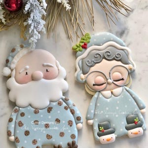 Peut inclure: Deux biscuits en sucre décorés, l'un représentant le Père Noël et l'autre une femme, tous deux portant un pyjama à pois. Les biscuits sont décorés de glaçage et ont une palette de couleurs bleu et blanc. Le texte "Cutter Designs By The Vintage Cookie Jar" est écrit sur les biscuits.