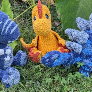May include: Three crocheted dinosaur toys on grass. A blue T-Rex with white details sits beside a yellow dinosaur with a red crest and feet. A blue Stegosaurus is also present.