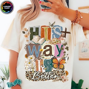May include: Cream-colored t-shirt with the text "HIS WAY IS BETTER" in a colorful, layered design. The design includes a cross, a dove, a butterfly, flowers, and an image of Jesus.