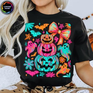 May include: Black t-shirt featuring a colorful Halloween design. The graphic includes stacked jack-o'-lanterns, ghosts, bats, flowers, and a large bow. The design uses bright neon colors like pink, orange, and teal.