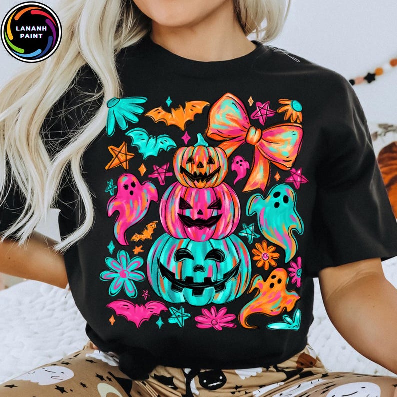 May include: Black t-shirt featuring a colorful Halloween design. The graphic includes stacked jack-o'-lanterns, ghosts, bats, flowers, and a large bow. The design uses bright neon colors like pink, orange, and teal.