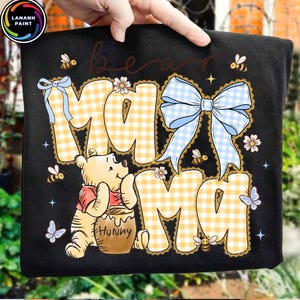 Pode incluir: Camiseta preta com um design do Ursinho Pooh. A camisa apresenta as palavras "Bear Mama" em um padr&atilde;o xadrez amarelo, um la&ccedil;o azul claro, abelhas e borboletas. Um urso de desenho animado est&aacute; segurando um pote de mel.