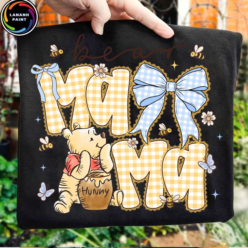 Pode incluir: Camiseta preta com um design do Ursinho Pooh. A camisa apresenta as palavras "Bear Mama" em um padr&atilde;o xadrez amarelo, um la&ccedil;o azul claro, abelhas e borboletas. Um urso de desenho animado est&aacute; segurando um pote de mel.