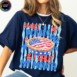 May include: Navy blue t-shirt with a graphic of a sweet potato pie decorated with an American flag design. The text reads "Sweet Potato Pie & I Shut My Mouth" in red and blue.