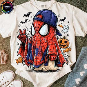May include: A white t-shirt featuring a graphic of a character wearing a red and blue spider-web patterned costume under a white sheet, topped with a blue baseball cap. The design includes a jack-o'-lantern, a ghost, bats, and candy.