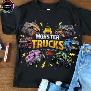 K&ouml;nnte beinhalten: Schwarzes T-Shirt mit einer farbenfrohen Grafik von Monstertrucks. Das Design zeigt verschiedene Monstertrucks in verschiedenen Farben, Blitze und den Text "MONSTER TRUCKS ARE MY JAM". Das Shirt wird mit blauen Jeans kombiniert.