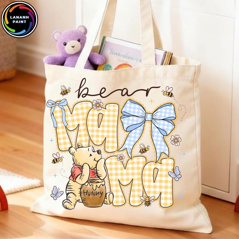 Pode incluir: Uma sacola bege com as palavras "bear mama" em uma fonte extravagante. As letras "MA" est&atilde;o em um padr&atilde;o xadrez amarelo e branco, com um la&ccedil;o azul. Winnie the Pooh &eacute; apresentado, junto com abelhas e borboletas.