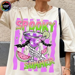 May include: Cream-colored t-shirt with a colorful graphic. The graphic features a spooky cocktail with ghost figures, bats, and watermelon slices. The words "SPOOKY SUMMER" are written in a dripping font. The shirt is a casual, oversized fit.