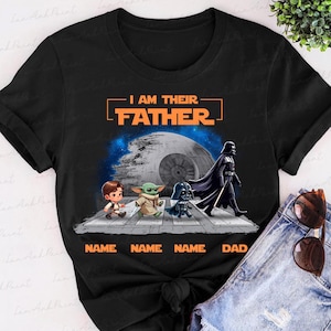 May include: Black t-shirt featuring a Star Wars-themed graphic. The design includes the text "I AM THEIR FATHER" above a depiction of characters crossing a street, with the Death Star in the background. The names are below the characters.