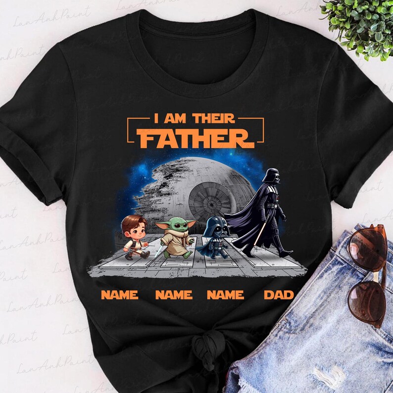 May include: Black t-shirt featuring a Star Wars-themed graphic. The design includes the text "I AM THEIR FATHER" above a depiction of characters crossing a street, with the Death Star in the background. The names are below the characters.
