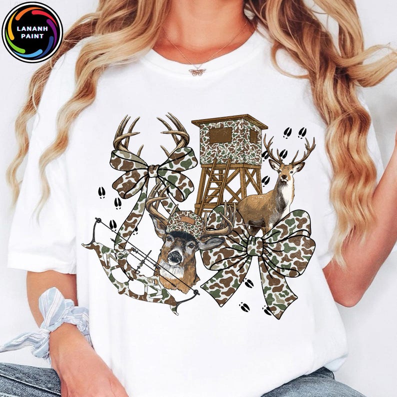 May include: White t-shirt featuring a deer-themed design. The graphic includes a deer, a hunting blind, a bow and arrow, and camouflage bows. The design incorporates brown, green, and white colors, with deer tracks scattered around the image.