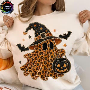 Può includere: Una felpa girocollo color crema con una grafica centrale raffigurante un fantasma che indossa un cappello da strega nero con stelle arancioni. Il fantasma ha una stampa leopardata e tiene in mano un secchiello a zucca nero, circondato da pipistrelli.
