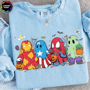 May include: A light blue crewneck sweatshirt featuring a graphic print of four cartoon characters dressed as ghosts for Halloween. The characters resemble Iron Man, Captain America, Spider-Man, and a green ghost, each holding a pumpkin candy bucket.
