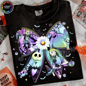 May include: Black t-shirt featuring a colorful graphic of characters from The Nightmare Before Christmas arranged in a bow shape. The design includes Jack Skellington, Sally, and Oogie Boogie, with a daisy in the center. The shirt has the text "LANANH PAINT" in a circle.