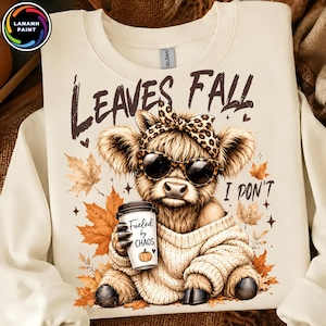May include: A cream-colored sweatshirt featuring a graphic of a fluffy Highland cow wearing leopard print sunglasses and a matching bow. The cow holds a coffee cup labeled "Fueled by CHAOS" with a pumpkin icon. Text reads "LEAVES FALL I DON'T".